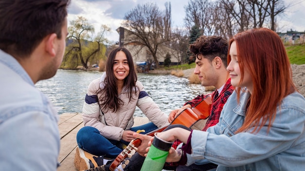 Quattro giovani amici cantano, riposano e suonano la chitarra vicino a un lago in un parco