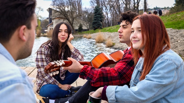 Quattro giovani amici cantano, riposano e suonano la chitarra vicino a un lago in un parco