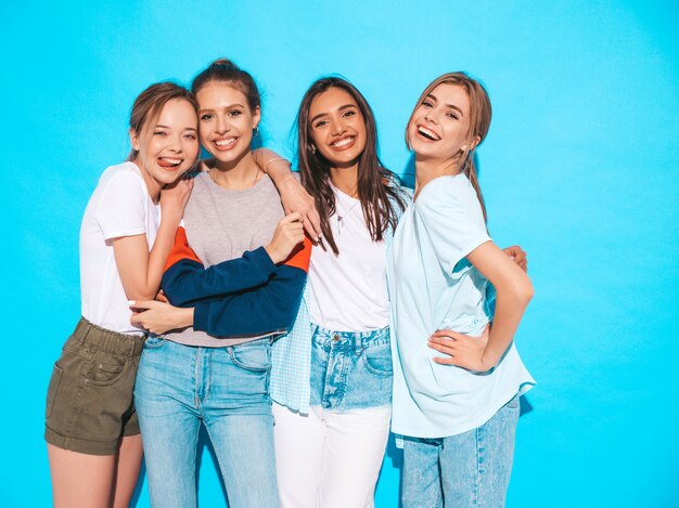 Quattro giovani belle ragazze sorridenti hipster in abiti estivi alla moda. Donne spensierate sexy che posano vicino alla parete blu in studio. Modelli positivi che si divertono e si abbracciano