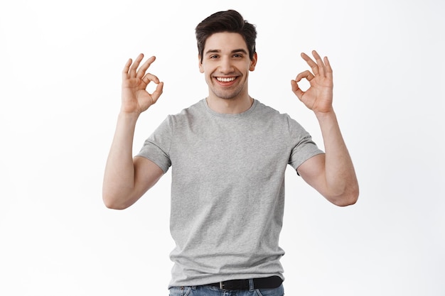 Questo va bene. Il bel ragazzo sorridente mostra segni ok, rassicura e dà garanzie tutto bene, loda un lavoro eccellente, in piedi su sfondo bianco