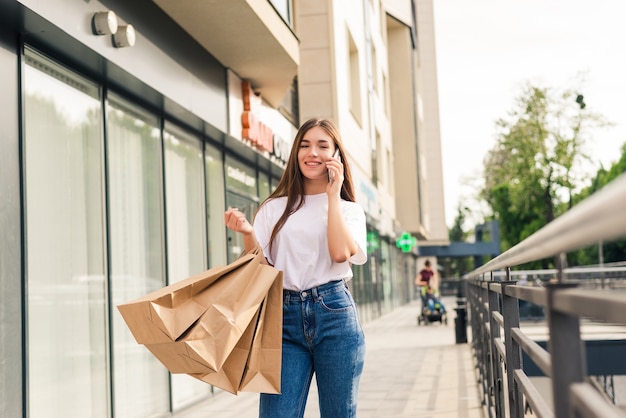 Raccontare ad un amico delle vendite. Bella giovane donna sorridente che tiene i sacchetti della spesa e parla al telefono cellulare mentre levandosi in piedi all'aperto