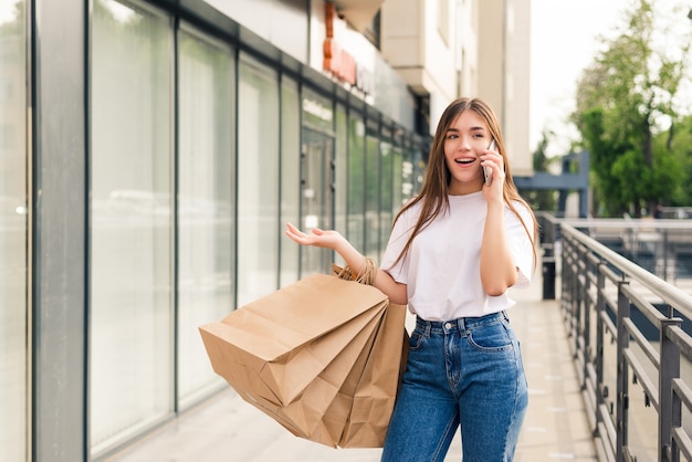 Raccontare ad un amico delle vendite. Bella giovane donna sorridente che tiene i sacchetti della spesa e parla al telefono cellulare mentre levandosi in piedi all'aperto