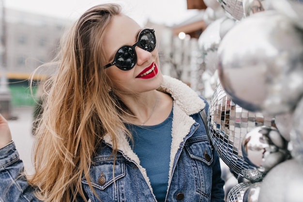 Raffinata donna caucasica indossa eleganti occhiali da sole neri sorridenti sulla strada urbana. Ritratto di bella ragazza bruna in giacca di jeans in posa in una buona giornata di primavera.