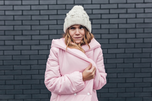 Raffinata giovane donna in abito invernale che fa facce buffe. Colpo all'aperto di spensierata ragazza caucasica in cappotto rosa e cappello lavorato a maglia.