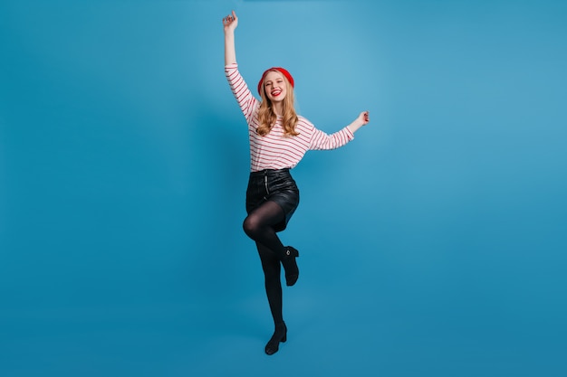 Raffinata ragazza bionda ballando e agitando le mani. magnifica donna francese in posa sulla parete blu.