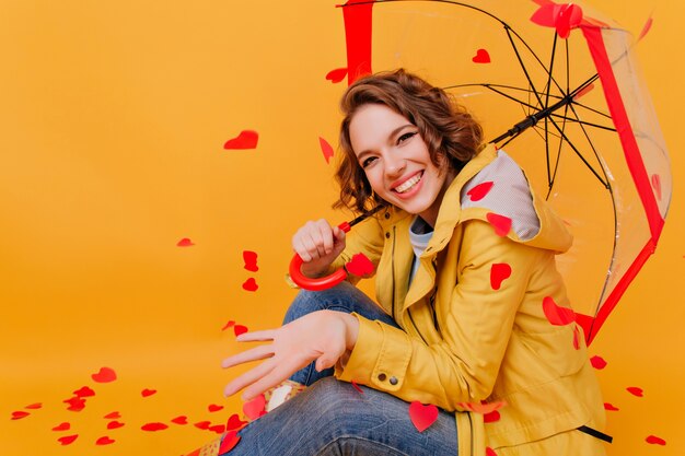 Raffinata ragazza che ride in giacca gialla alla moda in posa sotto l'ombrellone. Ritratto in studio di donna graziosa di buon umore seduto sul pavimento con i cuori sul muro.