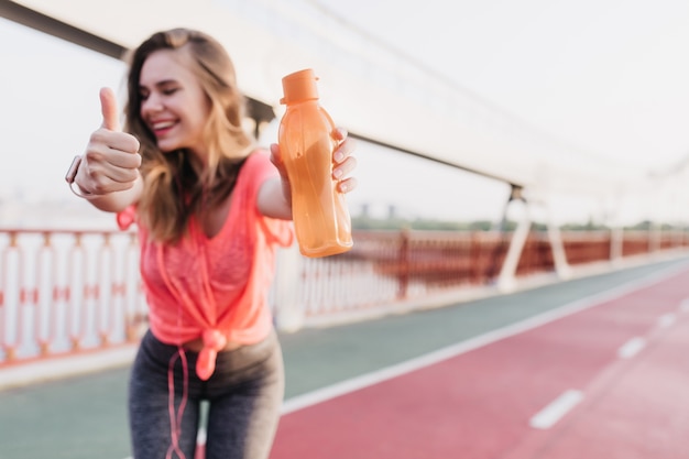 Raffinata ragazza in pantaloni sportivi grigi in posa con il sorriso. Ridendo graziosa donna in piedi con la bottiglia allo stadio.