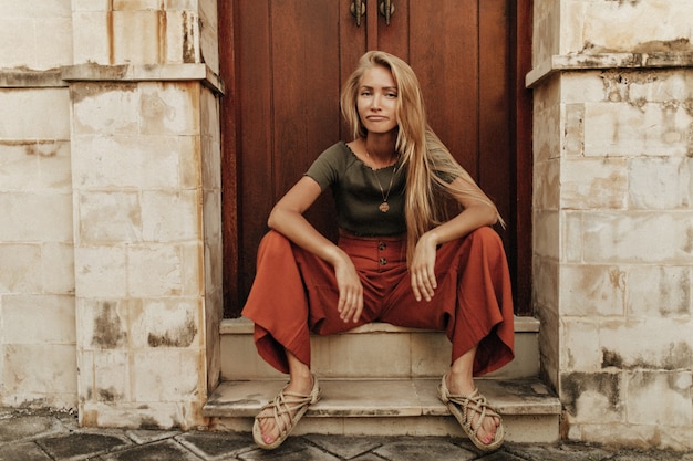 Raffreddare giovane bionda triste donna dai capelli lunghi in pantaloni rossi larghi e t-shirt cachi si siede in posa divertente vicino alla porta di legno e guarda in avanti