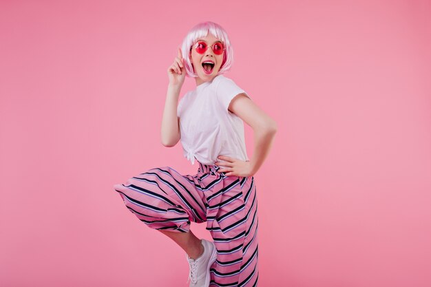 Ragazza abbastanza bianca in peruke lucido in posa in divertenti occhiali da sole rosa. Foto interna di graziosa signora in pantaloni alla moda che si gode il servizio fotografico e sorride