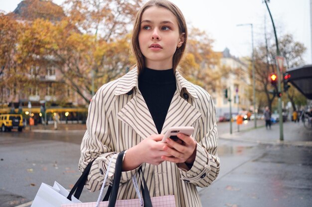 Ragazza abbastanza casual che cammina premurosamente per l'accogliente strada della città con borse della spesa e smartphone