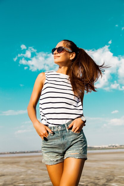 Ragazza abbastanza felice in pantaloncini di jeans e maglietta a strisce in posa con gli occhiali in spiaggia