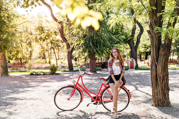 Ragazza abbastanza sottile che si siede sulla bici rossa. Donna alla moda gioconda che gode del fine settimana attivo.