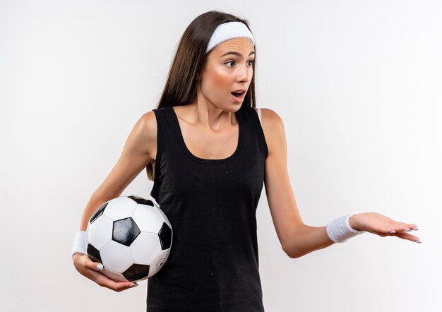 Ragazza abbastanza sportiva sorpresa che indossa la fascia e il wristband che tengono il pallone da calcio che esamina il lato e che mostra la mano vuota isolata su spazio bianco