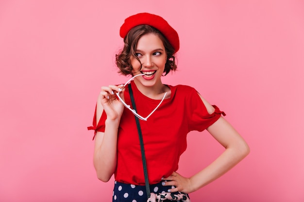 Ragazza accattivante con l'acconciatura riccia in posa con un sorriso giocoso. Tiro al coperto di spensierata signora francese in berretto rosso.