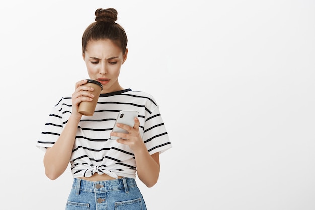 Ragazza accigliata confusa guardando lo schermo del telefono cellulare mentre si prepara un sorso di caffè dalla tazza