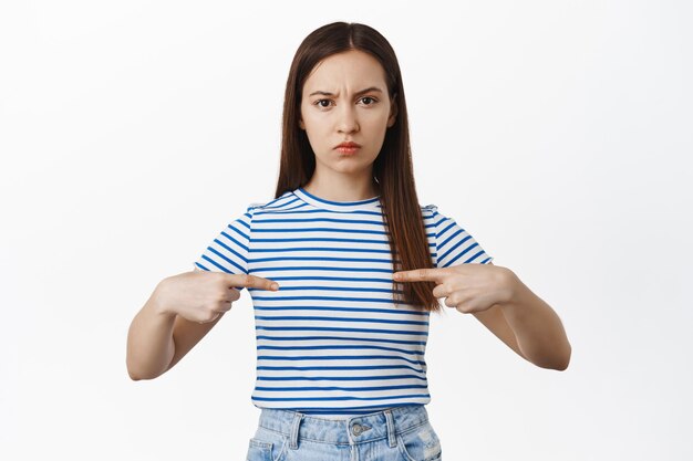 Ragazza accigliata sconvolta che punta il dito contro il logo al centro della maglietta, imbronciata delusa, antipatia, esprimere antipatia e disaccordo, in piedi su sfondo bianco