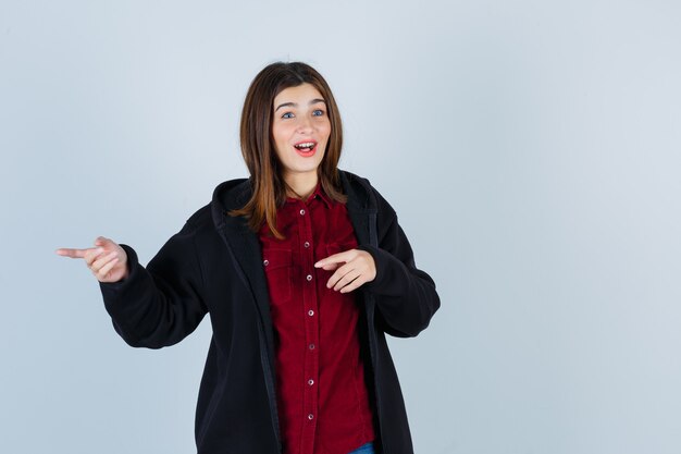 Ragazza adolescente che punta a sinistra in camicia, cappotto e guardando perplesso, vista frontale.