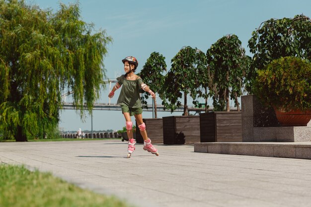 Ragazza adolescente in un casco impara a guidare sui pattini a rotelle tenendo un equilibrio o sui rollerblade e girare per la strada della città nella soleggiata giornata estiva