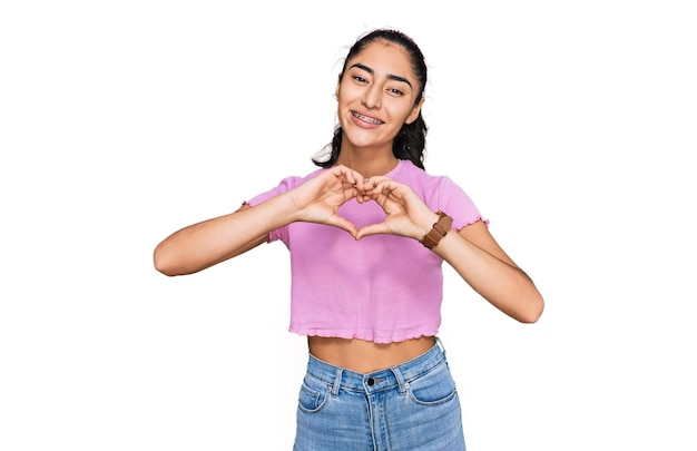 Ragazza adolescente ispanica con apparecchi ortodontici che indossa abiti casual sorridenti innamorati che fanno la forma del simbolo del cuore con le mani. concetto romantico.