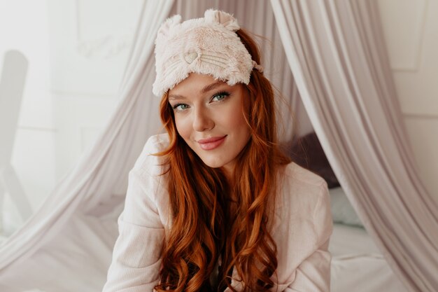 Ragazza adorabile vaga con capelli rossi ondulati lunghi con la mascherina di sonno che guarda l'obbiettivo nel letto al mattino