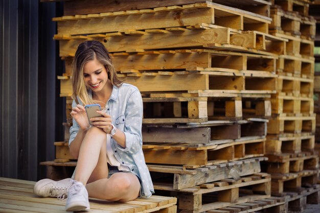 Ragazza affascinante che si siede su un pallet facendo uso dello Smart Phone