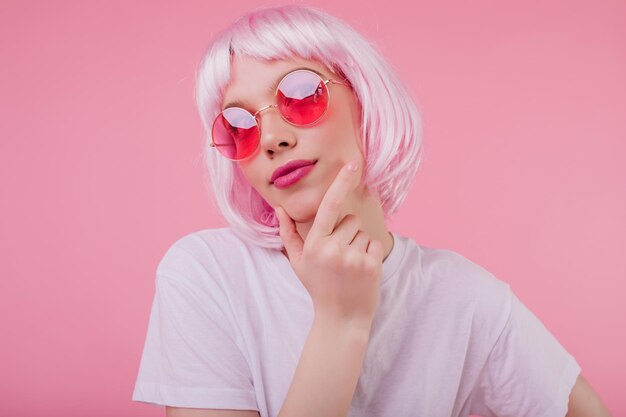 Ragazza affascinante pensierosa con i capelli rosa che guarda in alto mentre posa su sfondo pastello Donna europea spensierata in occhiali da sole e periwig agghiacciante durante il servizio fotografico in studio