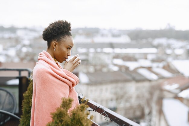 Ragazza africana sulla terrazza. Donna che beve il caffè in un plaid rosa. Signora in posa per una foto.