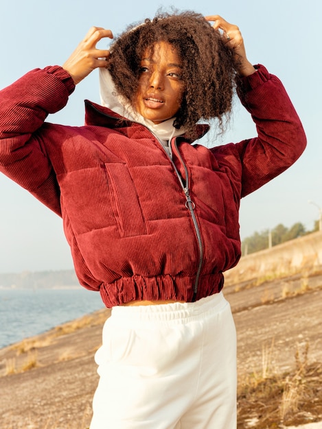 Ragazza afro in giacca rossa e pantaloni bianchi moderni, look alla moda. Brillante sorriso luminoso, corpo snello, capelli ricci voluminosi. Autunno freddo, vestiti caldi. All'aperto