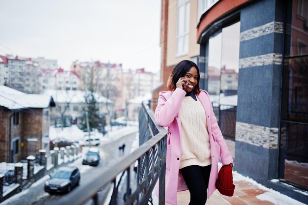 Ragazza afroamericana con cappello rosso e cappotto rosa in strada della città contro la costruzione in una giornata invernale con il telefono cellulare