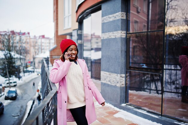 Ragazza afroamericana con cappello rosso e cappotto rosa in strada della città contro la costruzione in una giornata invernale con il telefono cellulare