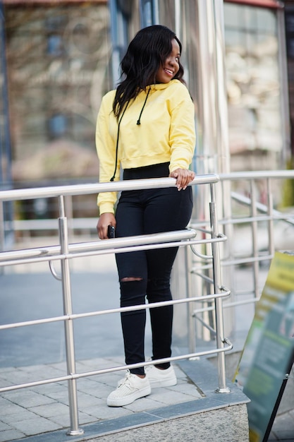 Ragazza afroamericana hipster che indossa una felpa con cappuccio gialla in posa in strada contro un edificio per uffici con finestre blu