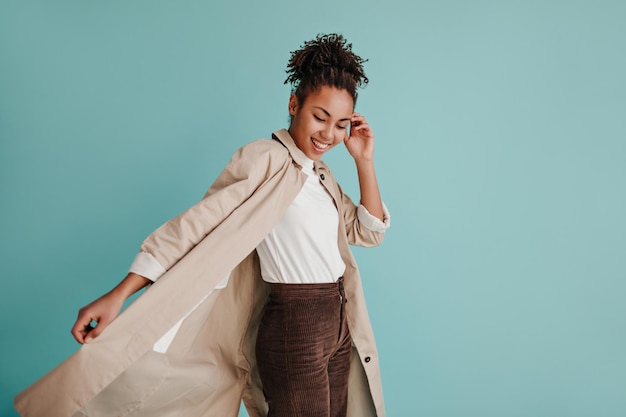 Ragazza afroamericana sicura in trench che guarda la fotocamera Donna nera allegra sorridente che posa su sfondo turchese