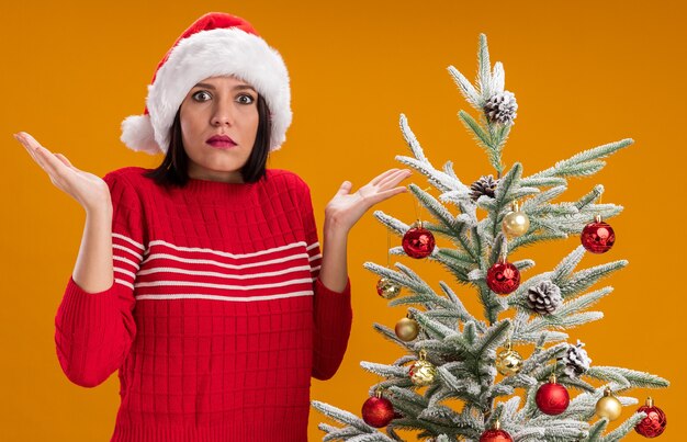 Ragazza all'oscuro che porta il cappello della Santa che sta vicino all'albero di Natale decorato che mostra le mani vuote isolate sulla parete arancione