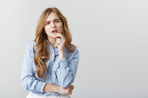 Ragazza alla moda che ha dubbi sul nuovo vestito. Studentessa incredula di bell'aspetto in camicia da colletto blu, toccandosi il labbro e fissando con espressione preoccupata, pensando al muro grigio