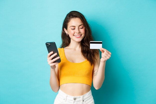 Ragazza alla moda con carta di credito in plastica sorridente, pagando per l'ordine online, facendo shopping su smartphone, in piedi su sfondo blu. Copia spazio