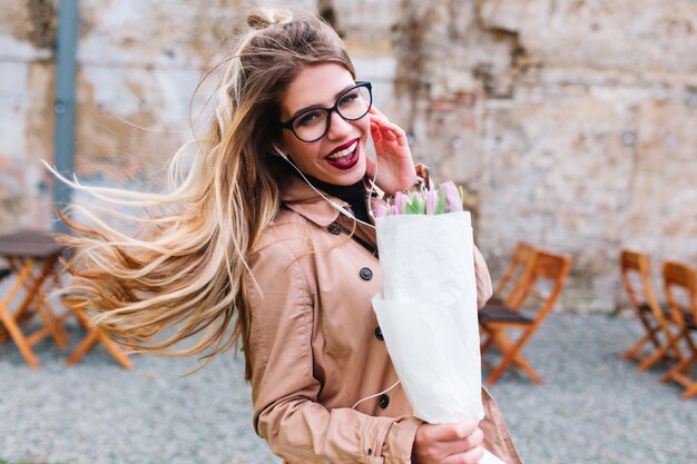 Ragazza alla moda con una bella acconciatura con gli occhiali scherza e ride portando il bouquet di tulipani. Adorabile giovane donna in giacca beige con capelli biondi in streaming sorridente sullo sfondo sfocato.