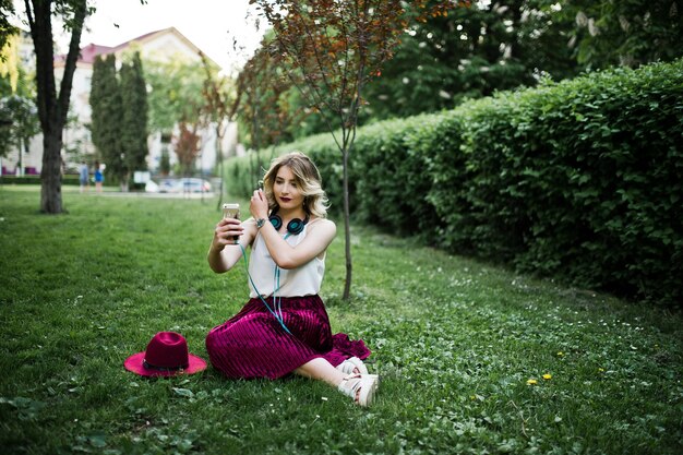 Ragazza alla moda e bella modella bionda in elegante velluto rosso gonna in velluto camicetta bianca e cappello seduto sull'erba verde al parco con telefono e auricolari Lei fa selfie