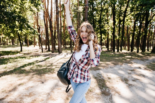 Ragazza alla moda emozionante divertendosi sulla foresta. Modello femminile allegro che esprime emozioni positive durante il viaggio.