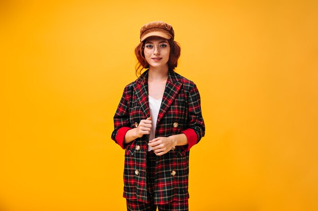 Ragazza alla moda in cappotto scozzese che posa sulla parete arancione