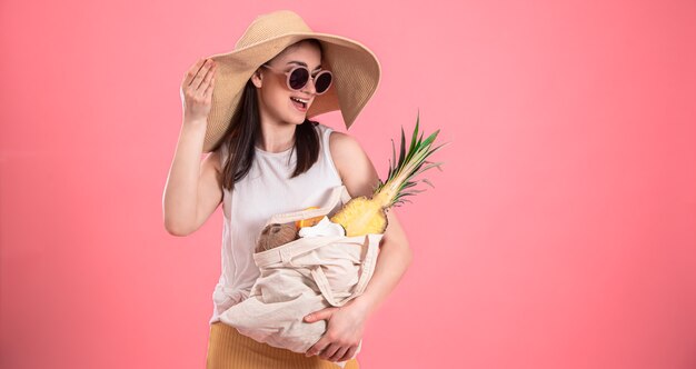 Ragazza alla moda in grande cappello e occhiali da sole sorride e tiene una borsa ecologica con frutti esotici