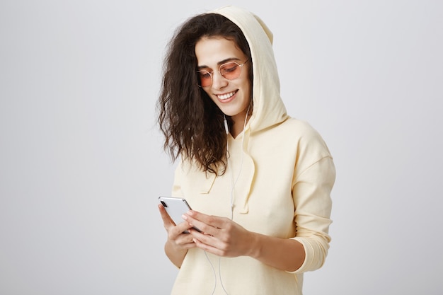 Ragazza alla moda in occhiali da sole che manda un sms tramite telefono cellulare e sorridente