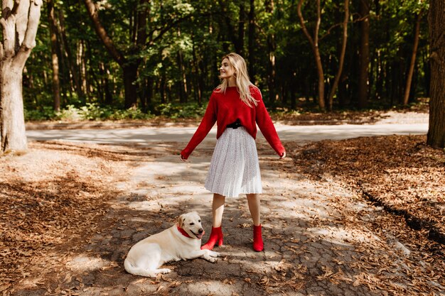 Ragazza alla moda in scarpe rosse divertendosi con il suo cane. Bionda allegra che sorride felicemente all'aperto.