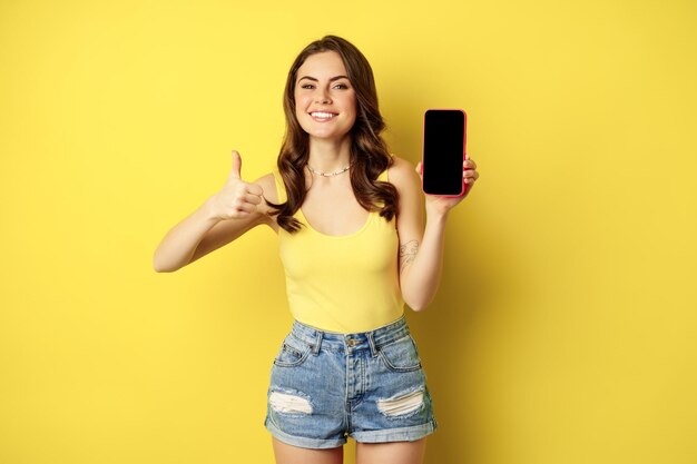 Ragazza alla moda sorridente felice che mostra i pollici in su e l'interfaccia dell'app per smartphone, il display del telefono cellulare, soddisfatta del sito Web, dell'applicazione per lo shopping online, in piedi su sfondo giallo.