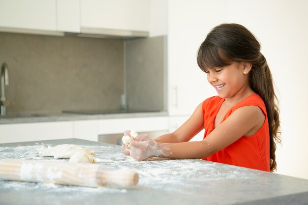 Ragazza allegra che impasta la pasta sul tavolo da cucina con farina disordinata e ridendo. Kid cuocere panini o torte da sola. Colpo medio. Concetto di cucina familiare