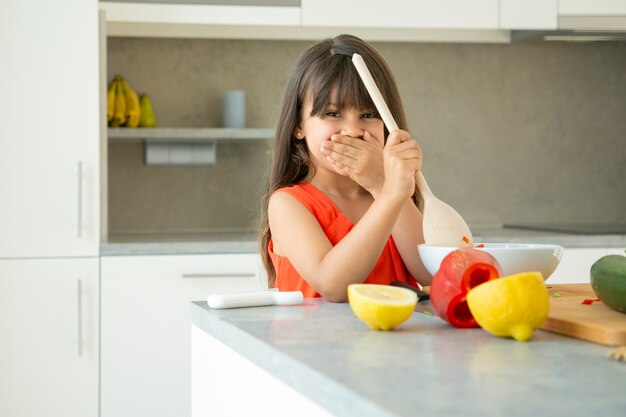 Ragazza allegra che lancia insalata nella ciotola con il grande cucchiaio di legno. Bambino sveglio che impara a cucinare le verdure per cena, in posa, sorridere alla telecamera. Imparare a cucinare il concetto