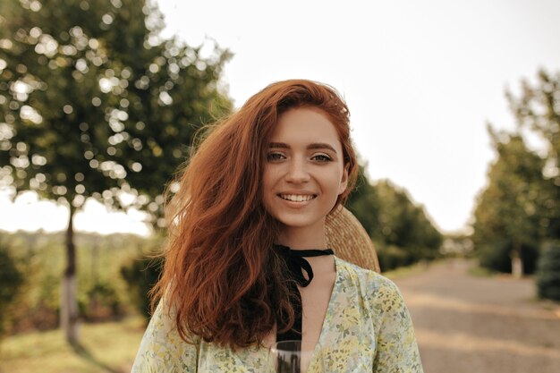 Ragazza allegra con acconciatura ondulata allo zenzero e benda nera sul collo in abito elegante estivo e cappello di paglia sorridente e guardando la parte anteriore all'aperto