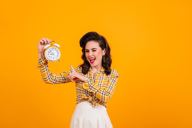 Ragazza allegra del pinup che mostra orologio. Studio shot di beata donna attraente isolata su sfondo giallo.