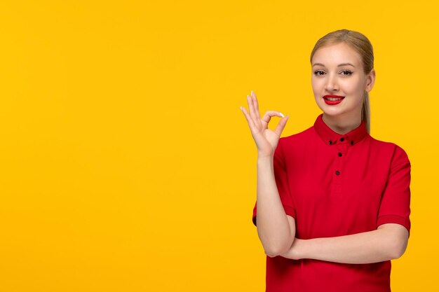 Ragazza allegra di giorno della camicia rossa che mostra il gesto giusto del segno in una camicia rossa su una priorità bassa gialla