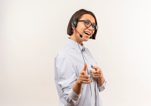 Ragazza allegra giovane call center con gli occhiali e auricolare in piedi nella vista di profilo che ti fa gesto davanti isolato su bianco