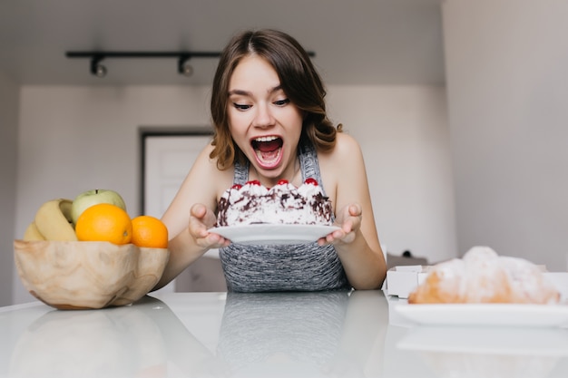 Ragazza allegra guardando la torta cremosa con desiderio. Tiro al coperto di raffinata giovane donna che scherza mentre mangia la torta.